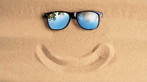Black sunglasses rest on sandy beach above a curved smile shape drawn in the sand, with palm trees reflected in the lenses against a bright sky