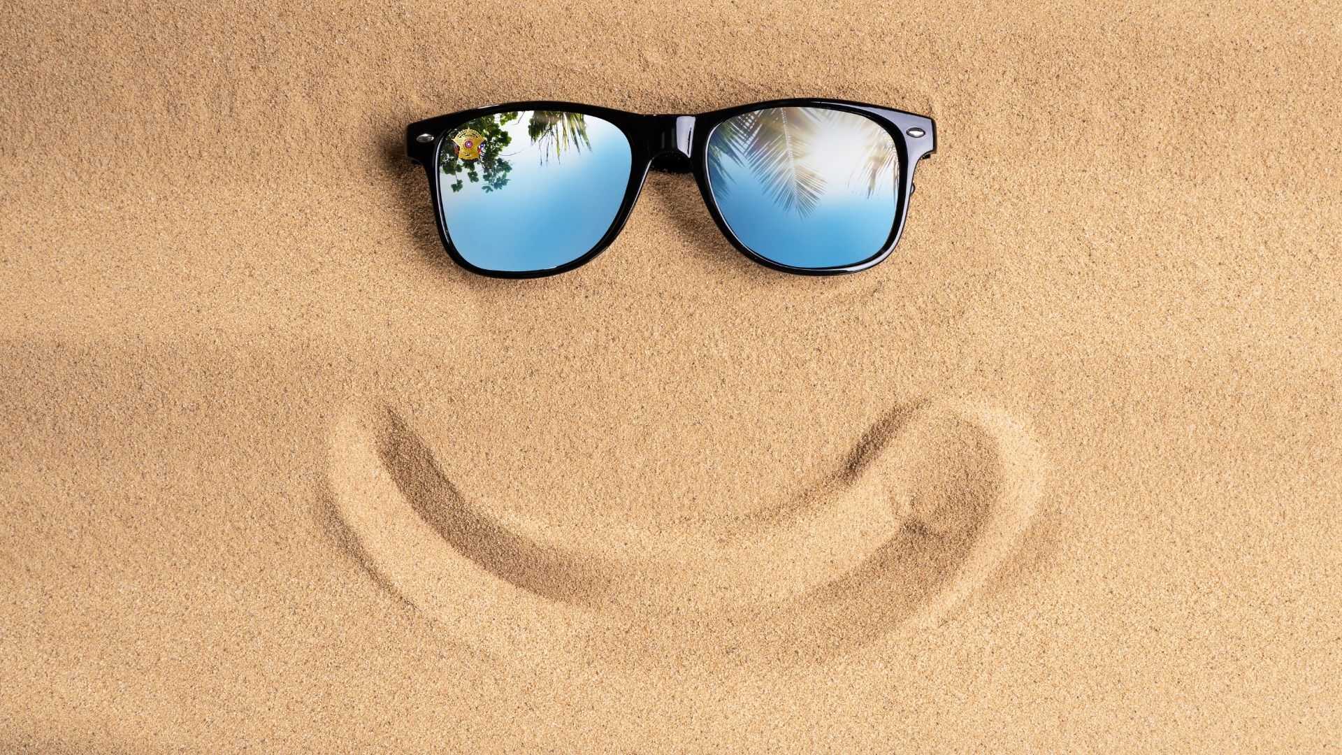 Black sunglasses rest on sandy beach above a curved smile shape drawn in the sand, with palm trees reflected in the lenses against a bright sky